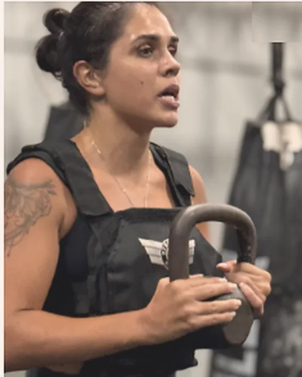 Ana Caroline Morais treinando na academia, com colete de peso e kettlebell nas mãos.